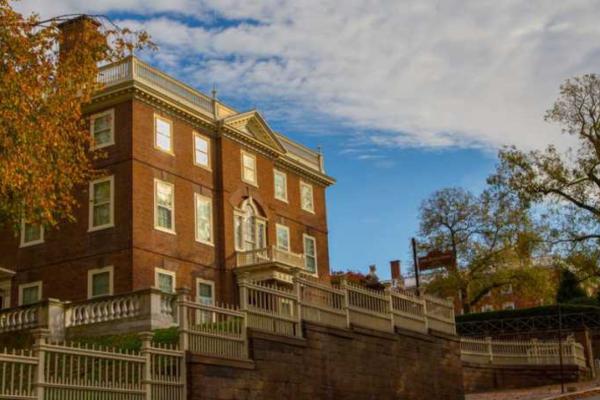 A large brick building with white trim and classical details stands behind a tall white fence, framed by autumn trees under a partly cloudy sky.