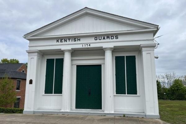 A small white historic building with four columns and green shutters stands at a roadside; the sign above the door reads “Kentish Guards 1774.”