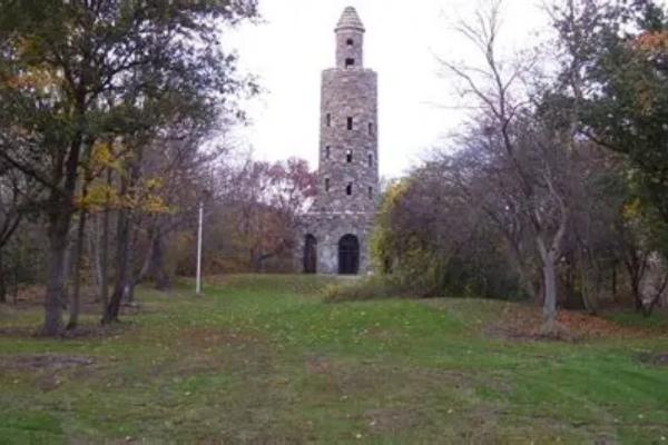 A tall cylindrical stone tower with small windows and an arched entrance stands in a grassy clearing surrounded by trees showing early autumn color.