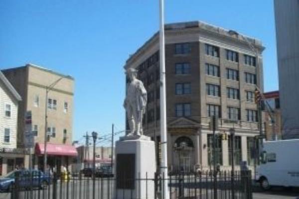 A colonial era militia statue enclosed by a circular black fence stands in a small plaza with an American flag behind it, surrounded by multi‑story city buildings under a bright blue sky.