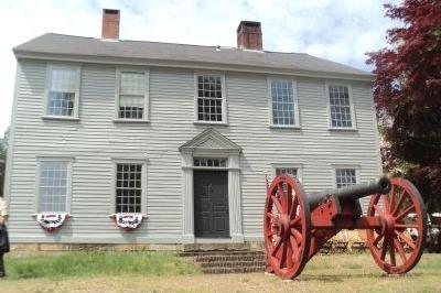 Colonial era 2-story manor house with 9 windows trimmed in white, with 2 chimneys and a red cannon in the front yard.