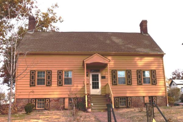 A two‑story yellow colonial house with dark brown trim and twin brick chimneys stands behind a small front porch and brick walkway.