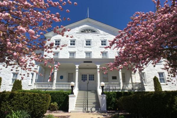 A white colonial‑style building with a central staircase and double doors is framed by bright pink flowering trees, with an American flag near the entrance and a clear blue sky overhead.