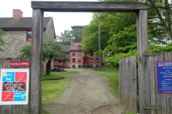 A wooden entrance gate stands at the Old Barracks Museum, built from two tall posts and a horizontal beam.  Beyond the gate, a gravel path leads to historic stone and brick buildings with red trim, surrounded by trees and greenery.