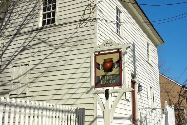 A white historic building with a stone foundation and an exterior stairway stands behind a sign reading “Potter’s Tavern 1773” topped with a brown pot illustration.
