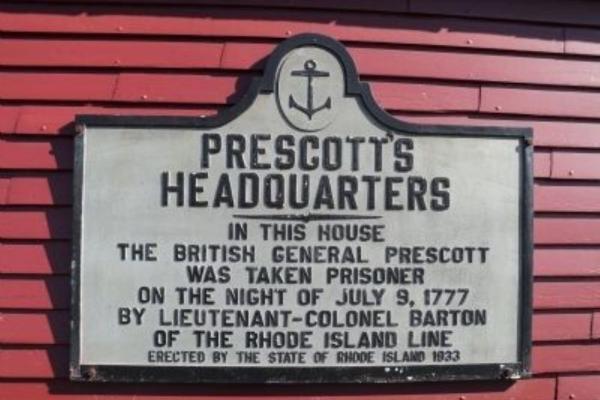 A rectangular historical marker to British General Prescott mounted on a red wooden wall.