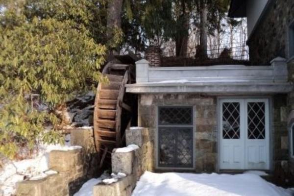 A rustic stone mill with a large wooden water wheel stands in the snow among evergreen trees.