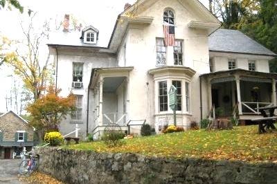 A large light‑colored house with multiple gables, a stone wall in front, and an American flag above the porch sits among autumn trees with fallen leaves.