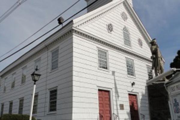A white wooden church with a tall clock‑tower steeple and red doors stands behind a stone‑bordered lawn with a lamppost and statue.
