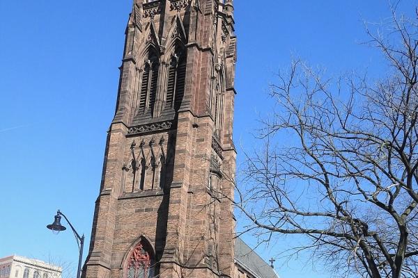 A tall brown stone Gothic‑style church tower with pointed arches and ornate spires rises above trees under a bright blue sky.