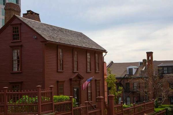 A reddish‑brown historic wooden house with tall narrow windows and an American flag at the entrance stands behind a matching rod iron fence.