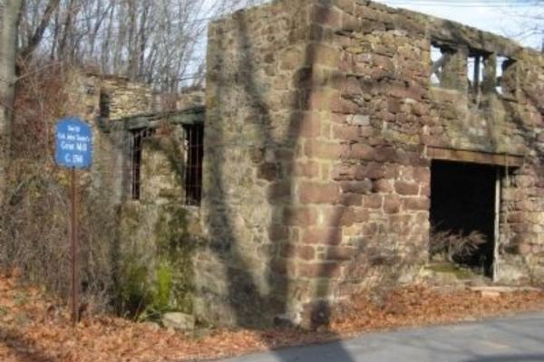 The stone ruins of an old grist mill stand among leafless trees, with a blue historical marker identifying the site beside the weathered walls.