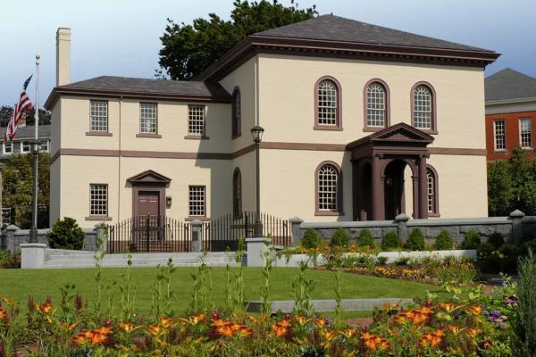 A beige building with brown trim and a columned portico stands behind a garden of bright flowers and neatly trimmed shrubs, bordered by a wrought‑iron fence and stone posts.