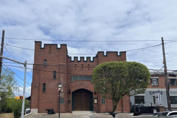 A small fortress like brick building sits on a street corner, with arched wooden doors, battlements, and a nearby cannon.