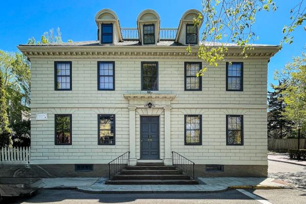 A light‑colored historic building with dark shutters and a central double‑door entrance sits on a corner lot, featuring three dormer windows on the roof and a short staircase leading up from the sidewalk.