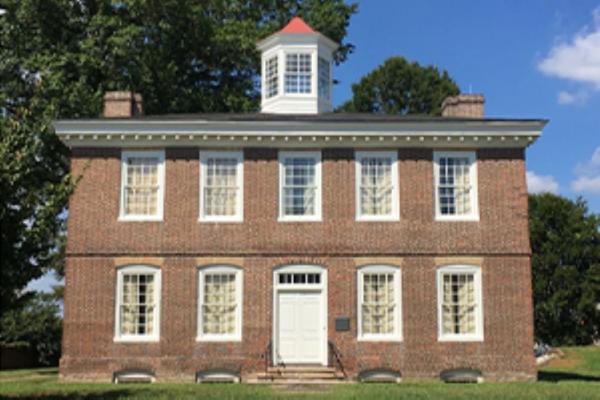 A two‑story brick Federal style building with white‑trimmed windows and a central cupola stands on a grassy lawn beneath a bright blue sky.