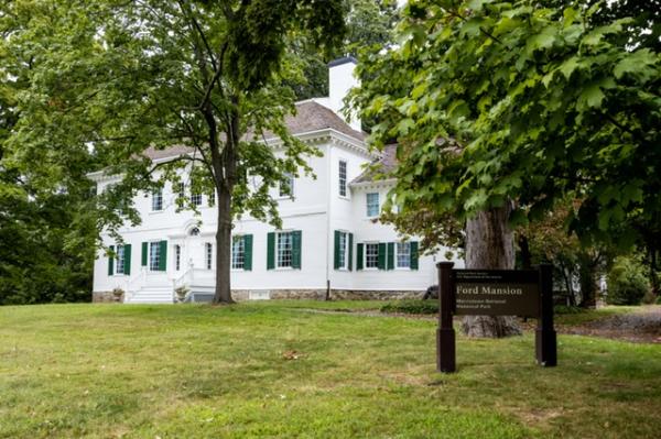 A white two-story colonial house with dark green shutters on the bottom floor stands in the background surrounded by trees. A brown sign stands in the foreground with the title, "Ford Mansion" legible.