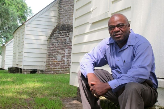 Joe McGill of the Slave Dwelling Project sitting in front of a historic cabin.