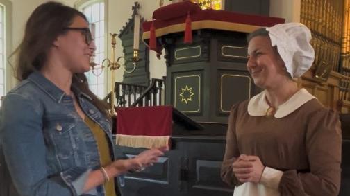 One woman in modern clothes gestures in conversation to another women dressed in historical clothing as Abigail Adams. They are standing inside historic St. John's Church. 