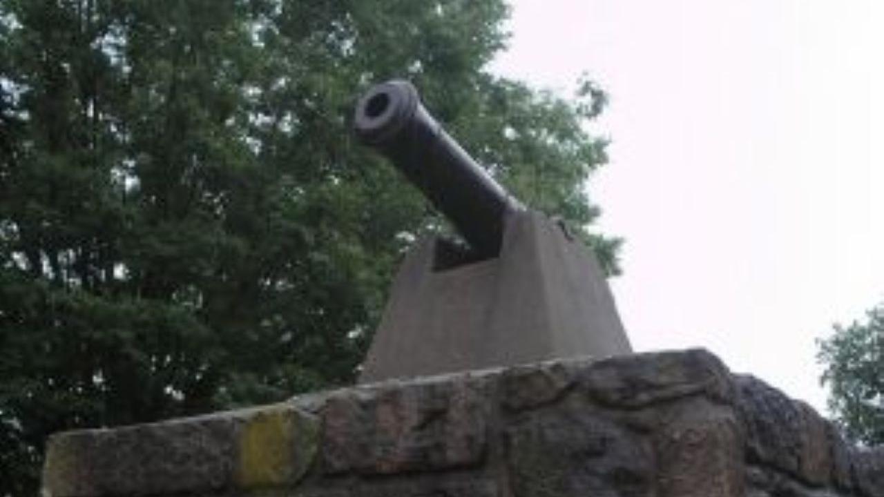 A black cannon sits atop a small stone structure with a glass window, surrounded by green trees under an overcast sky.