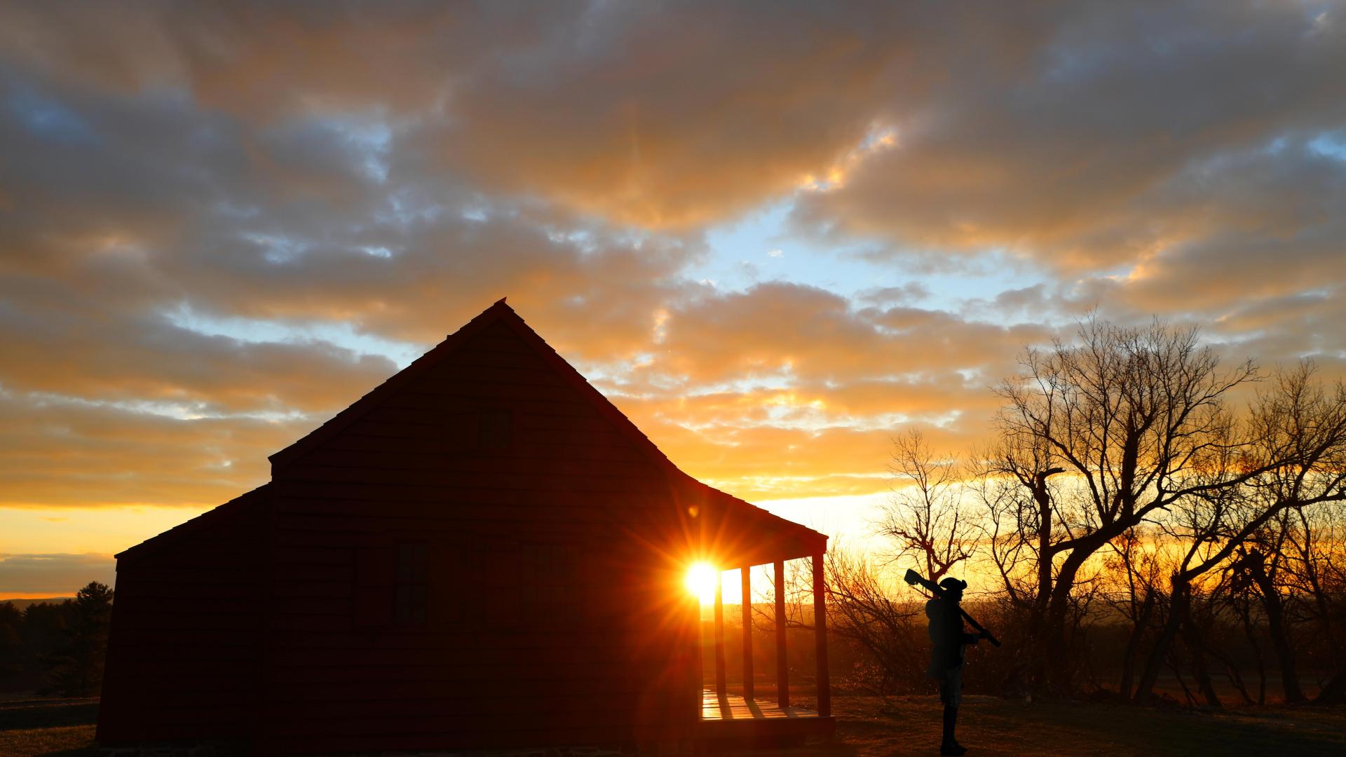 A man wearing 18th-century clothing carrying a musket and a farmhouse are silhouetted by sunrise.