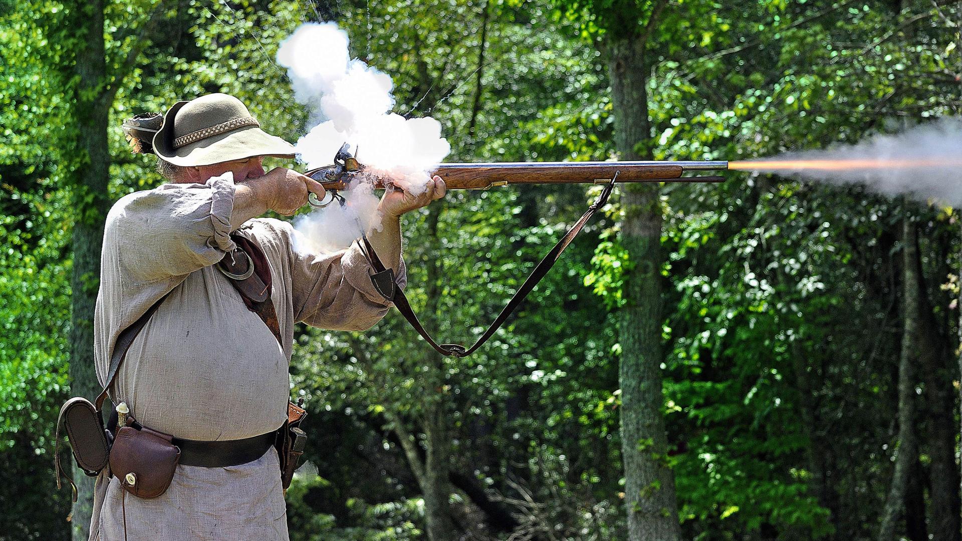 Smoke puffs up from the vent and a flame shoots out of the muzzle as a militiaman fires his musket.