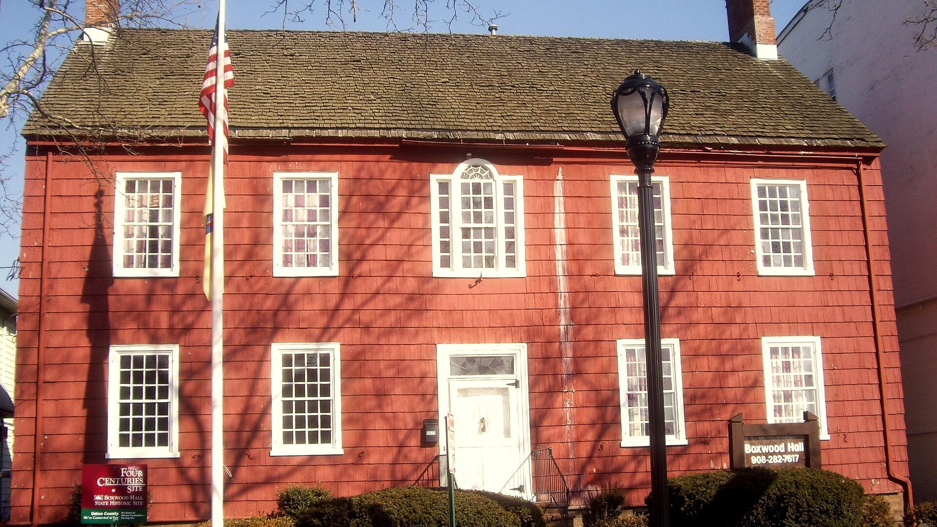 A red historic house with white trim and twin brick chimneys stands behind a flagpole and walkway lined with bushes.