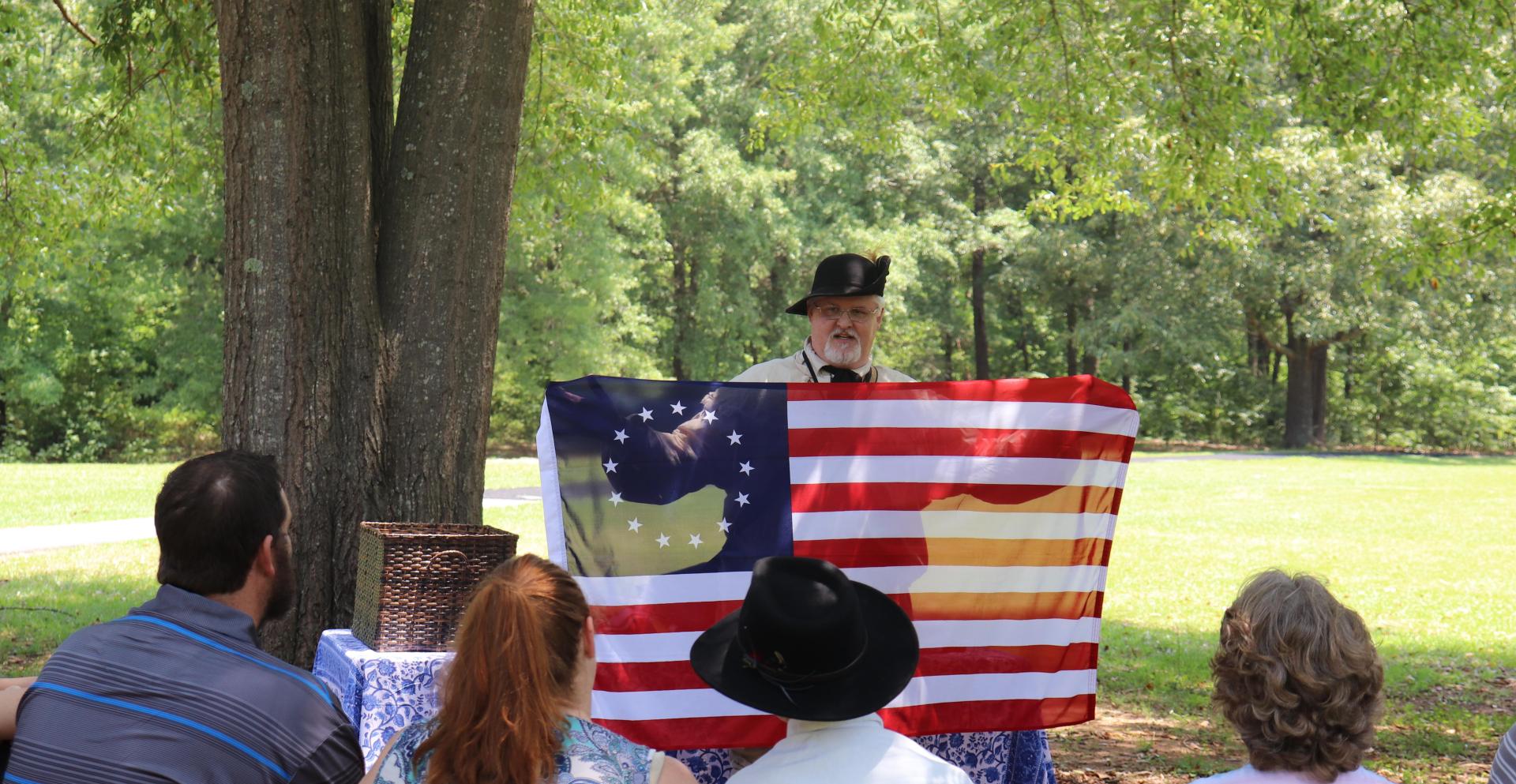 Volunteer dressed as a 18th century militia person holds a flag for a group of visitors.