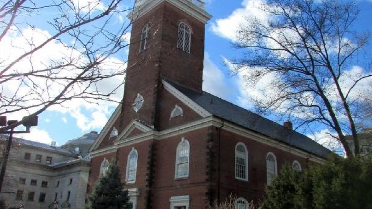 A tall brick church with a white clock‑tower steeple rises above a fenced grassy area with trees under a bright blue sky.