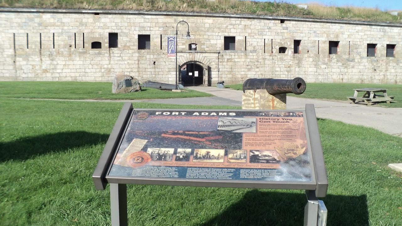A stone fort wall with an arched entrance rises behind a grassy area where an informational sign, an old cannon, and a picnic table sit under a bright blue sky.