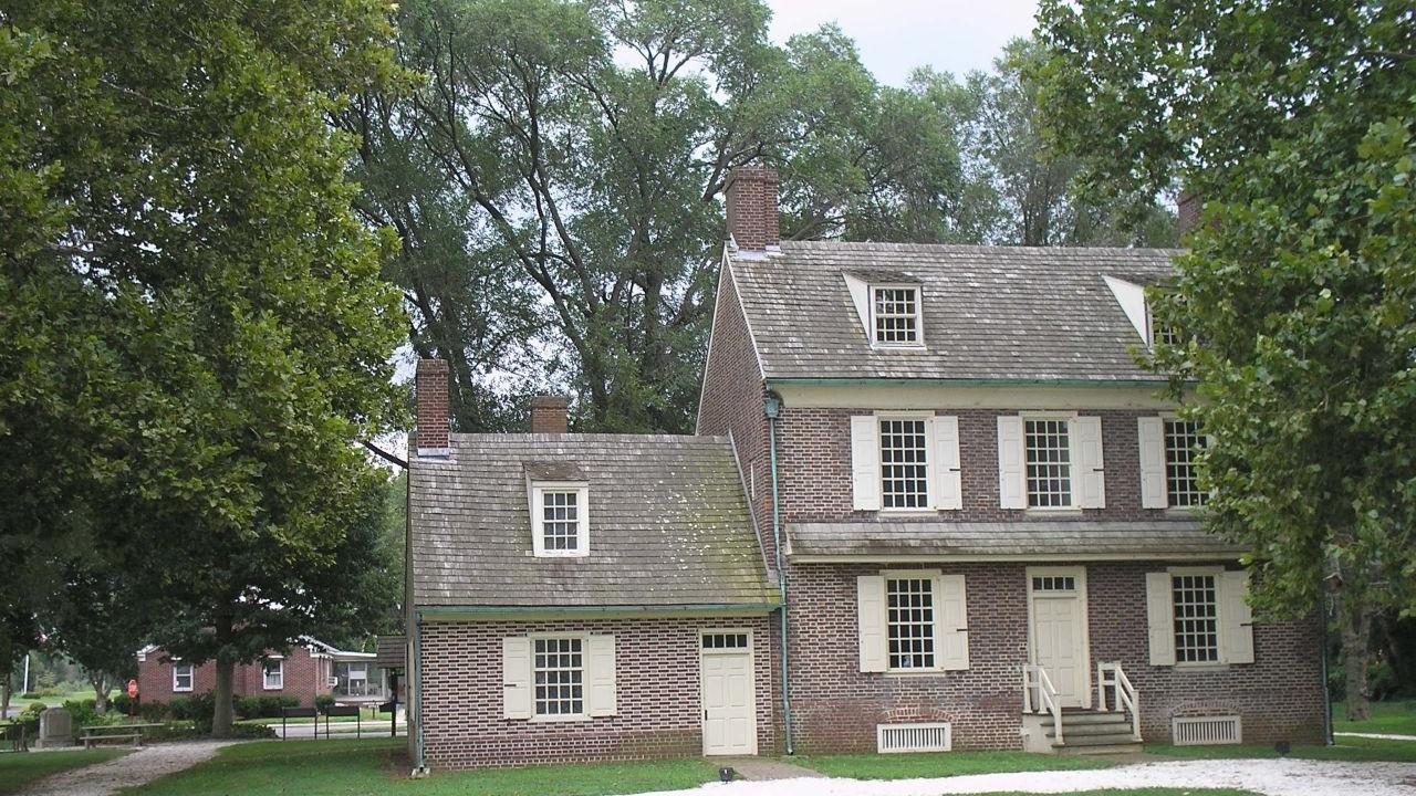 A brick colonial‑style house with cream shutters and multiple chimneys stands amid green grass and tall trees.