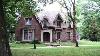 A large brick Gothic‑style house with steep gables and tall narrow windows stands among mature trees on a grassy lawn.