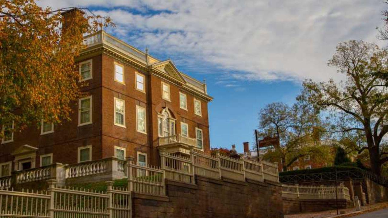 A large brick building with white trim and classical details stands behind a tall white fence, framed by autumn trees under a partly cloudy sky.