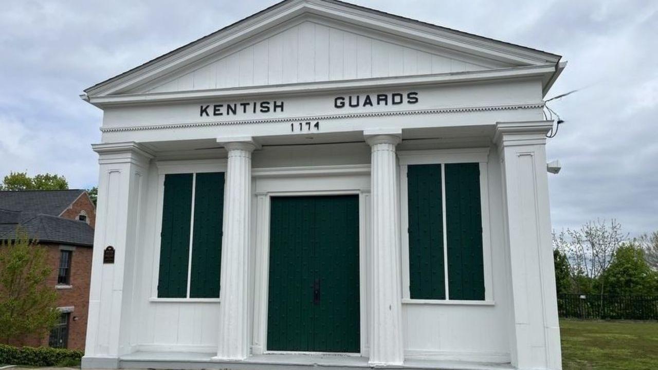 A small white historic building with four columns and green shutters stands at a roadside; the sign above the door reads “Kentish Guards 1774.”