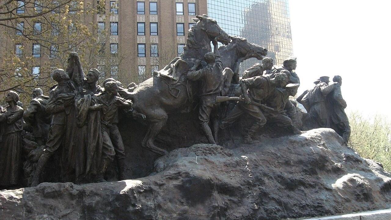 A large bronze monument shows soldiers and civilians in dramatic motion, with horses and figures emerging from a rugged base in an outdoor urban park.