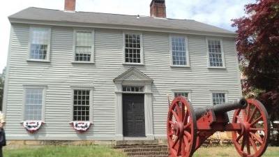 Colonial era 2-story manor house with 9 windows trimmed in white, with 2 chimneys and a red cannon in the front yard.