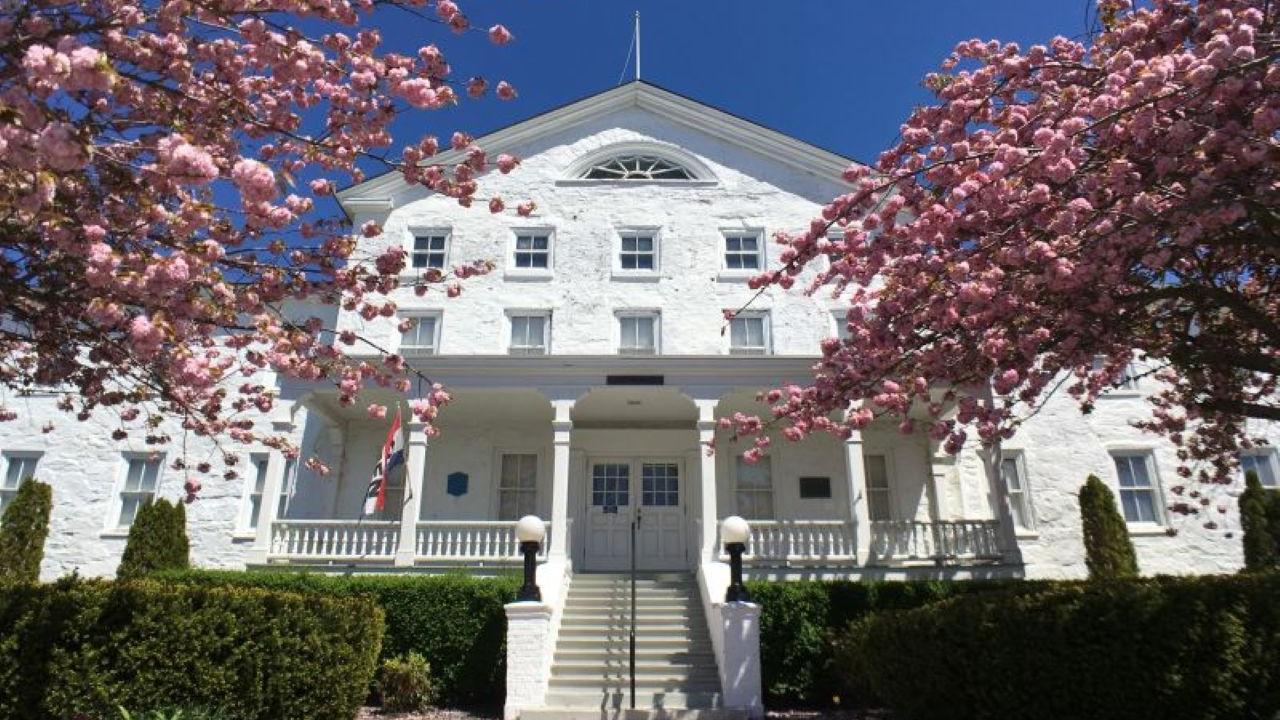 A white colonial‑style building with a central staircase and double doors is framed by bright pink flowering trees, with an American flag near the entrance and a clear blue sky overhead.