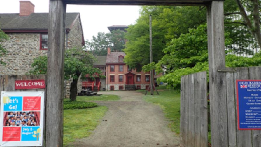 A wooden entrance gate stands at the Old Barracks Museum, built from two tall posts and a horizontal beam.  Beyond the gate, a gravel path leads to historic stone and brick buildings with red trim, surrounded by trees and greenery.