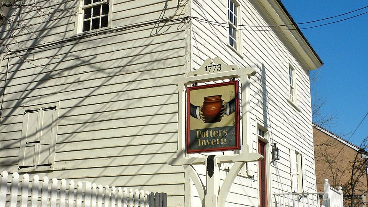 A white historic building with a stone foundation and an exterior stairway stands behind a sign reading “Potter’s Tavern 1773” topped with a brown pot illustration.