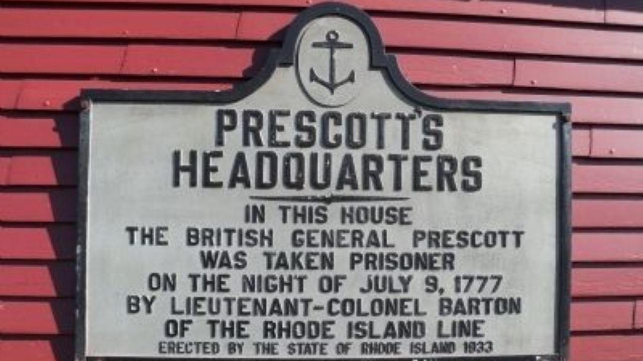 A rectangular historical marker to British General Prescott mounted on a red wooden wall.