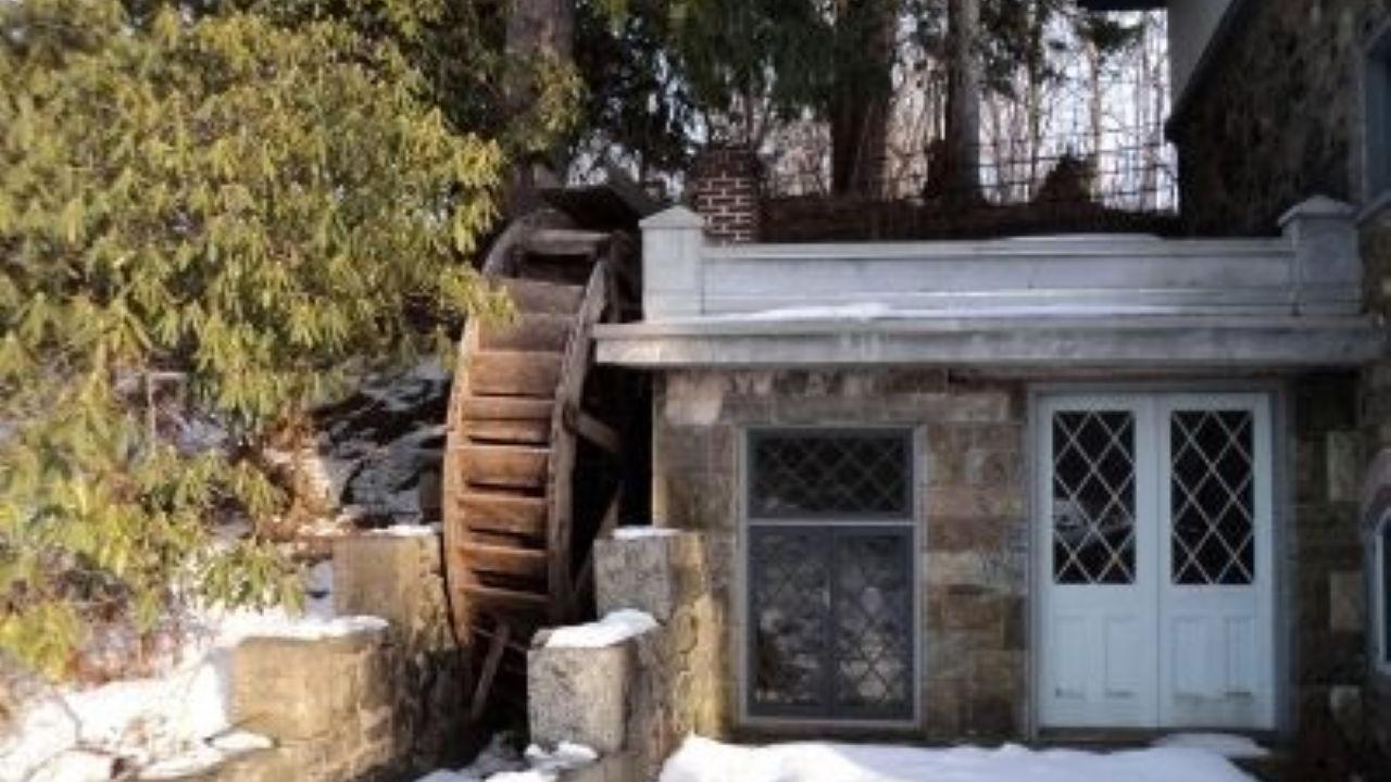 A rustic stone mill with a large wooden water wheel stands in the snow among evergreen trees.