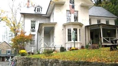 A large light‑colored house with multiple gables, a stone wall in front, and an American flag above the porch sits among autumn trees with fallen leaves.