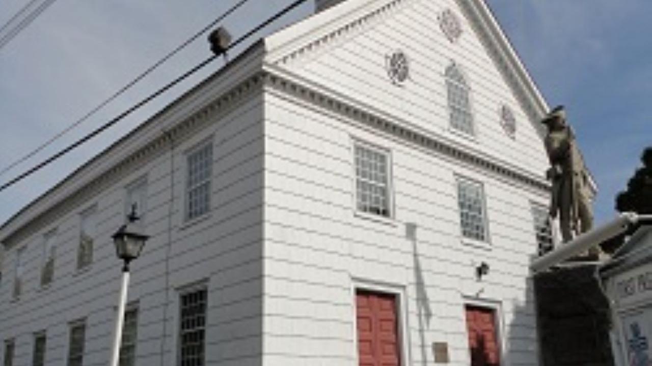 A white wooden church with a tall clock‑tower steeple and red doors stands behind a stone‑bordered lawn with a lamppost and statue.