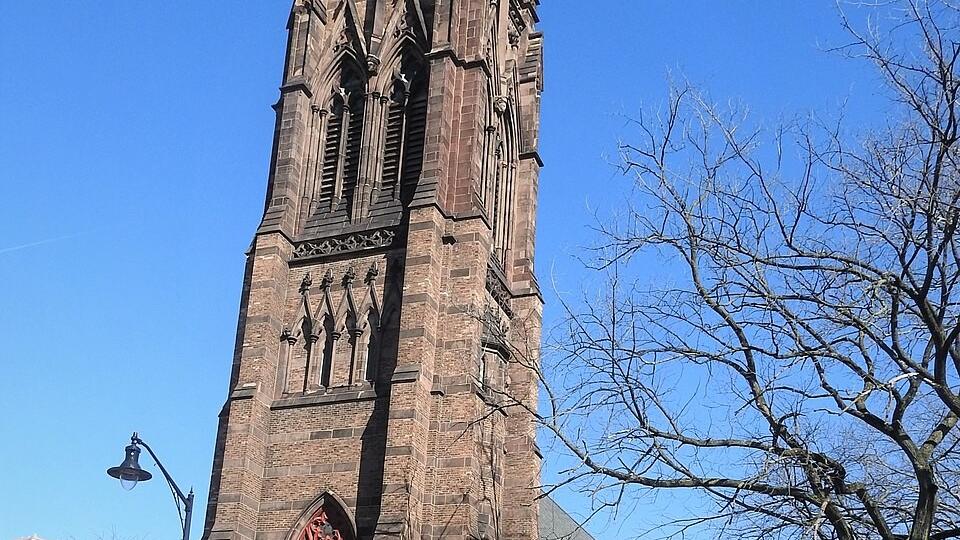 A tall brown stone Gothic‑style church tower with pointed arches and ornate spires rises above trees under a bright blue sky.