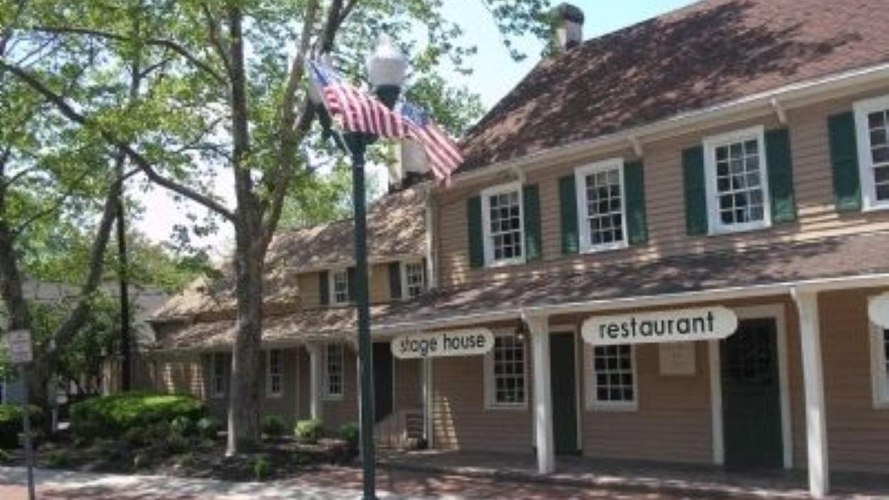 A tan historic building with green shutters and a columned porch stands along a brick sidewalk, shaded by large trees and marked by a streetlamp flying an American flag.