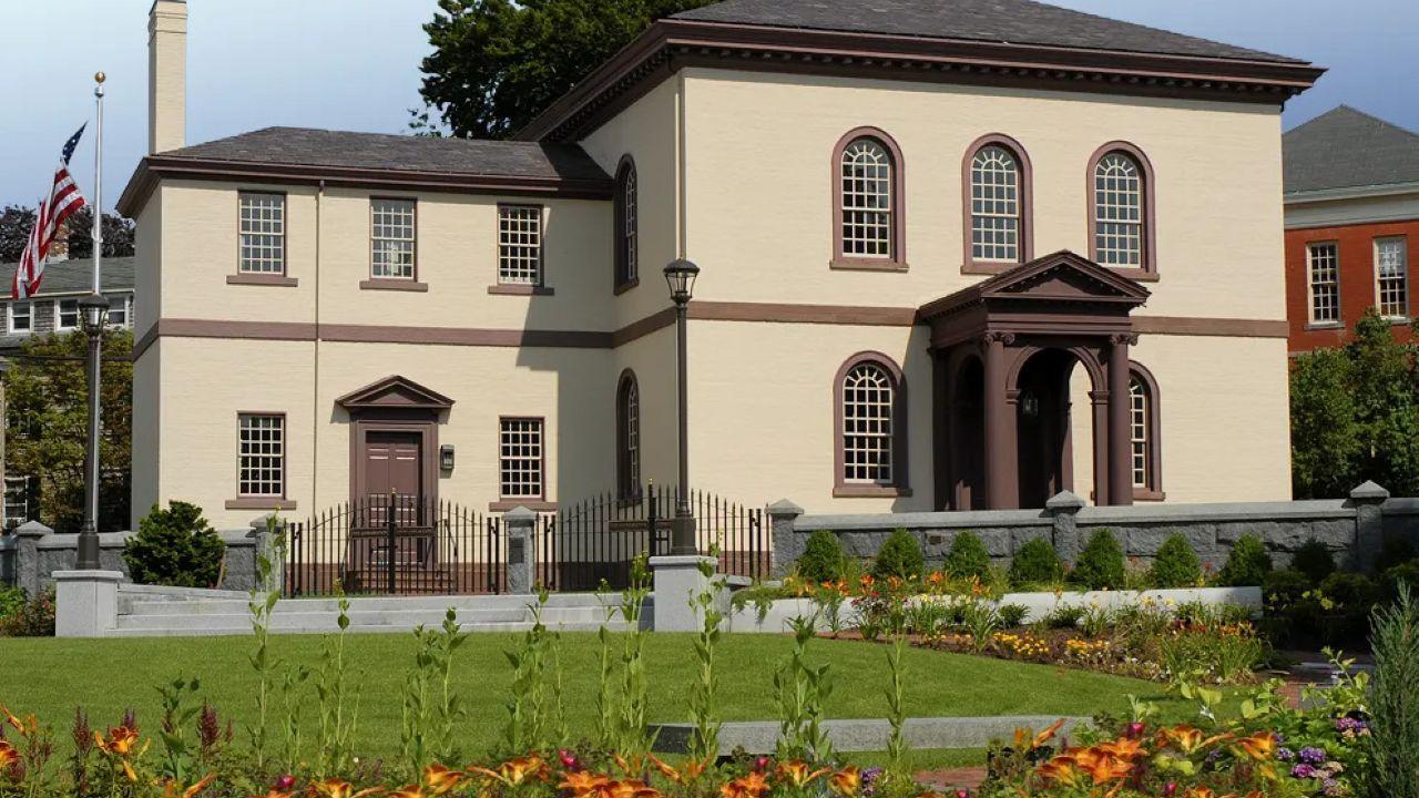 A beige building with brown trim and a columned portico stands behind a garden of bright flowers and neatly trimmed shrubs, bordered by a wrought‑iron fence and stone posts.