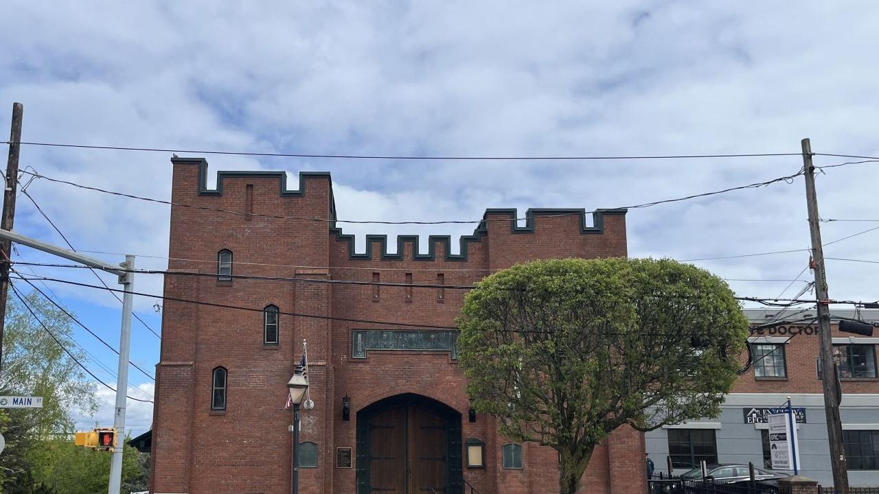 A small fortress like brick building sits on a street corner, with arched wooden doors, battlements, and a nearby cannon.