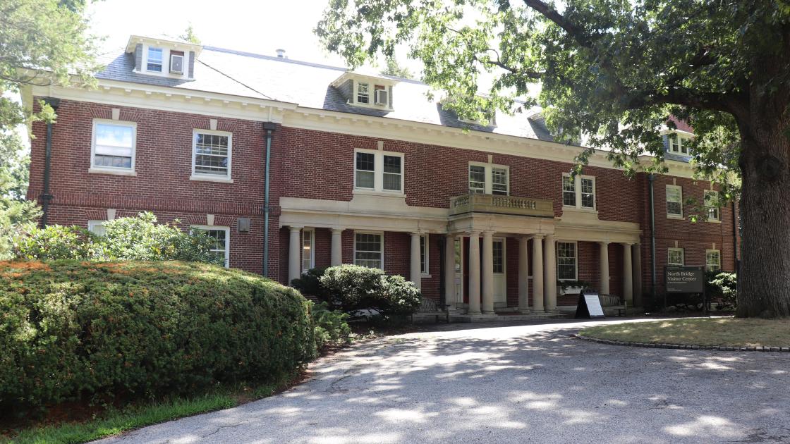 A three story brick mansion house with paved walkway leading to door. Green bushes flank both sides.