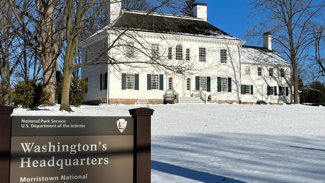 A white colonial building with snow in the foreground and a blue sky and trees in the background