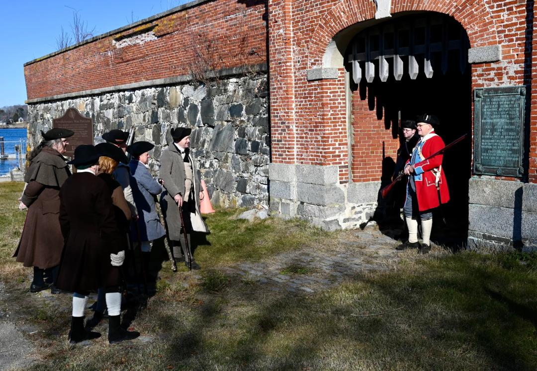 A group of patriot soldiers stand outside the gate of a fort guarded by a reenactor wearing a British uniform. 